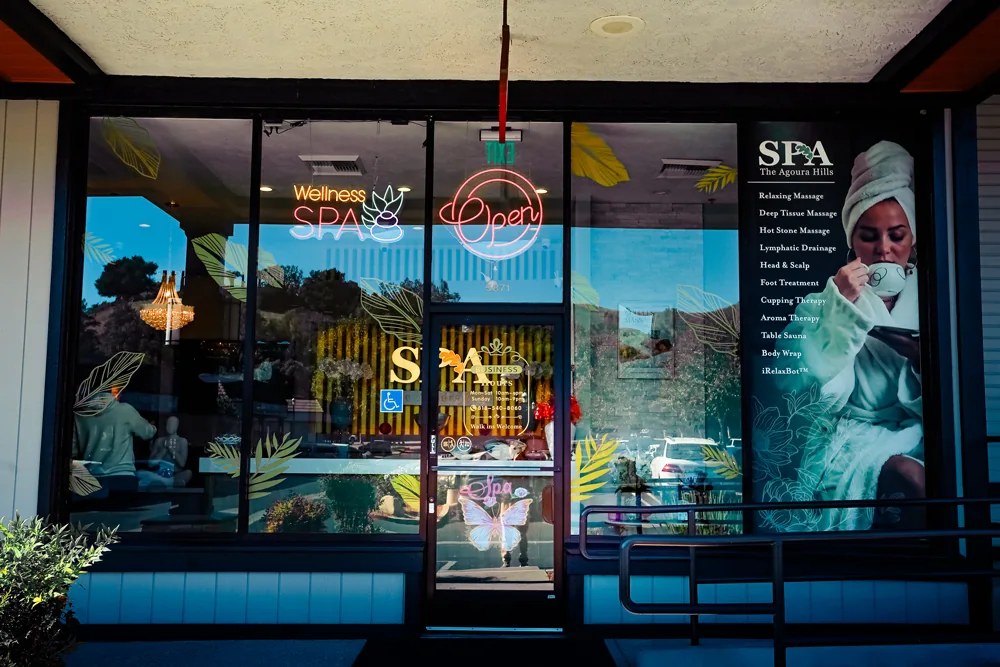 Glass entrance of The Agoura Hills Spa with neon Wellness SPA and Open signs and a service-menu poster