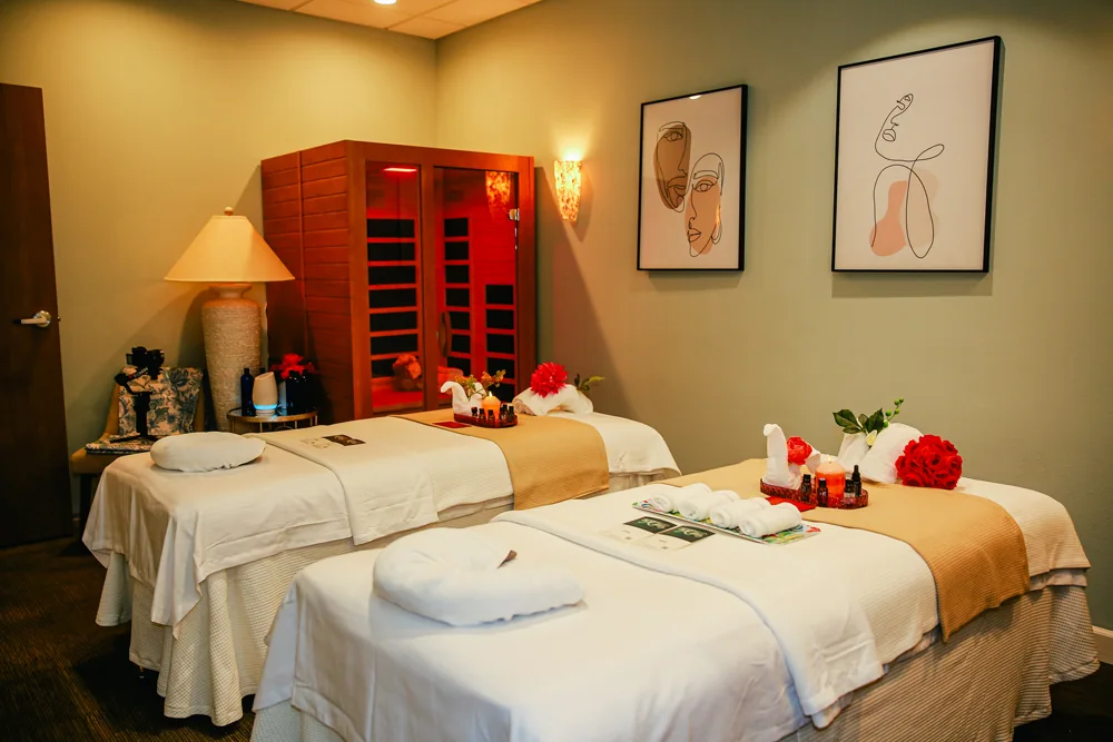 Couples room with two side-by-side massage tables, towel swans, roses, and a red-lit infrared sauna cabin